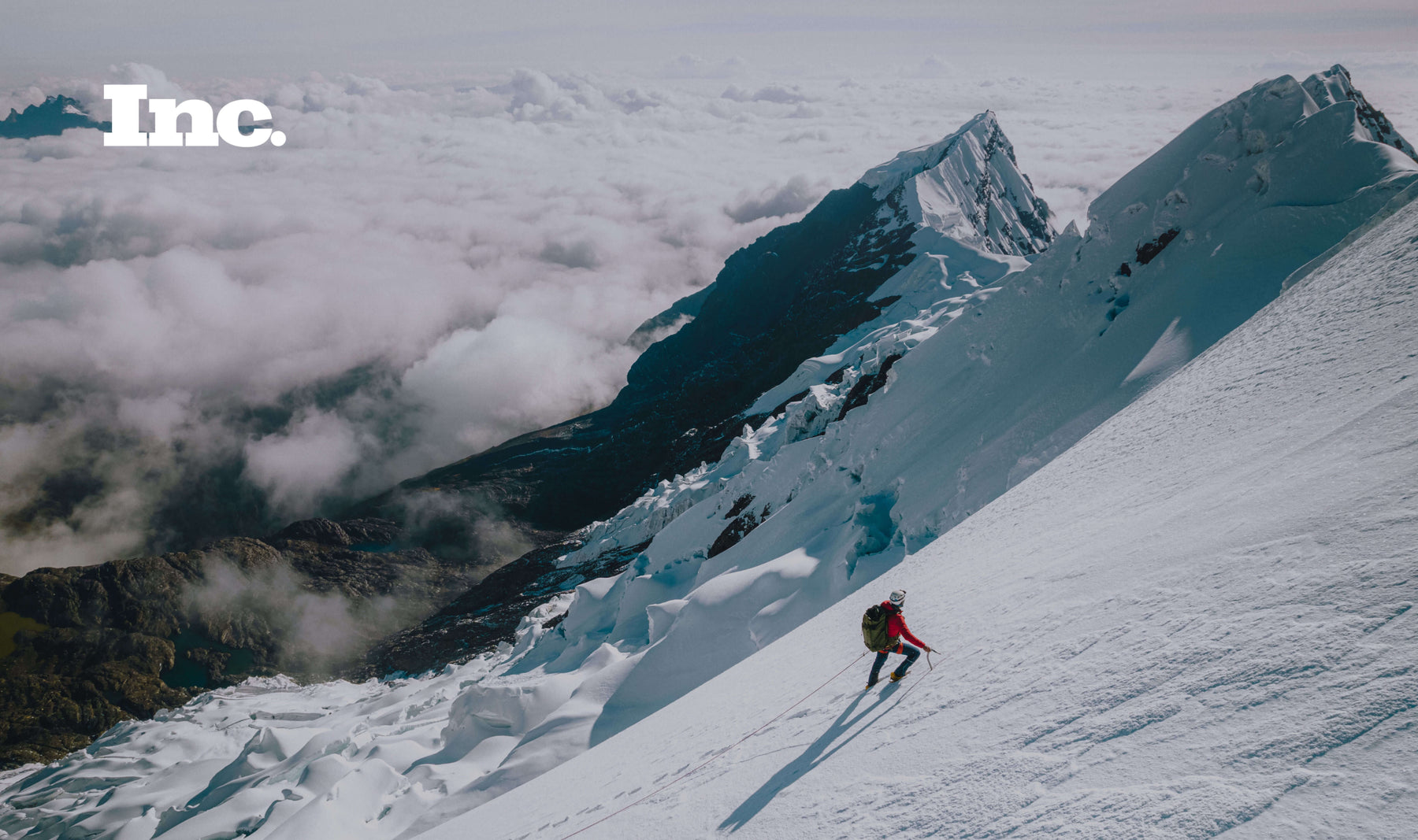 A man wearing a red Pakafill puffer jacket and a helmet summiting a snowy mountain