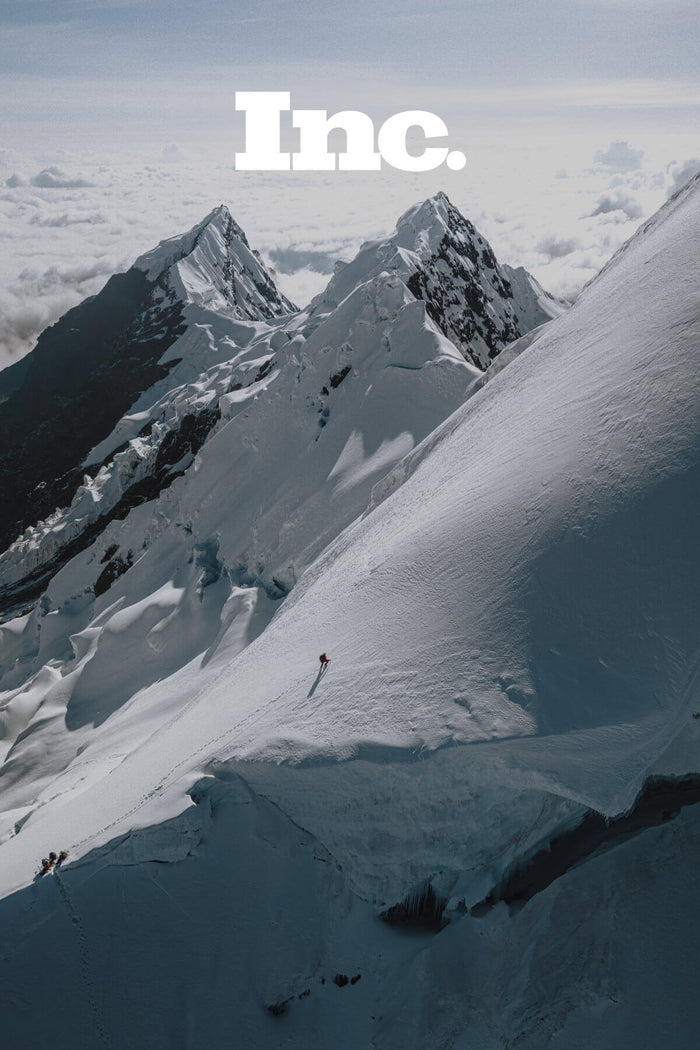 A man wearing a red Pakafill puffer jacket and a white helmet summiting a snowy mountain