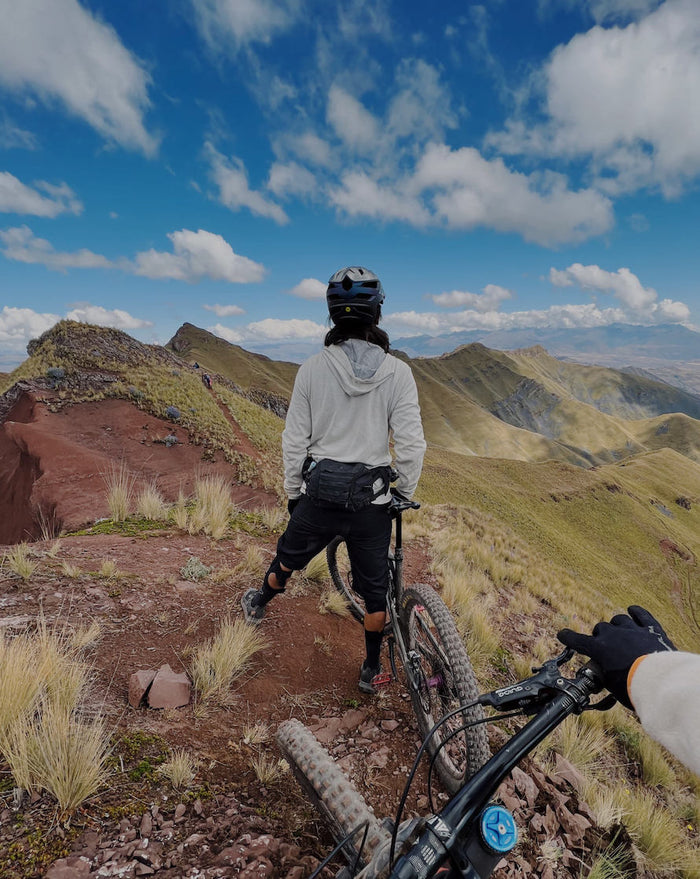 Man wearing alpaca hoodie standing on ridge of mountain next to mountain bike 