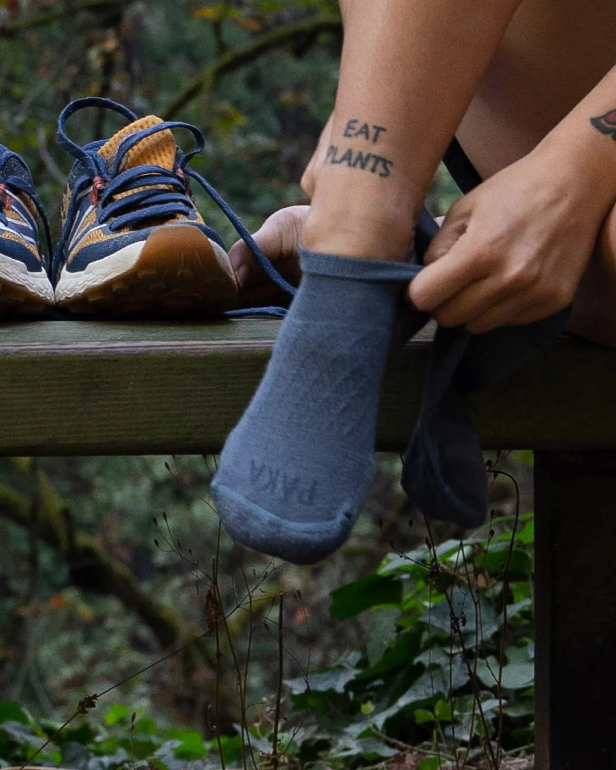 Leg of Woman with “Eat Plants” Tattoo Pulling on an Indigo Blue Alpaca Wool Ankle Sock Next to Running Shoes – PAKA Apparel