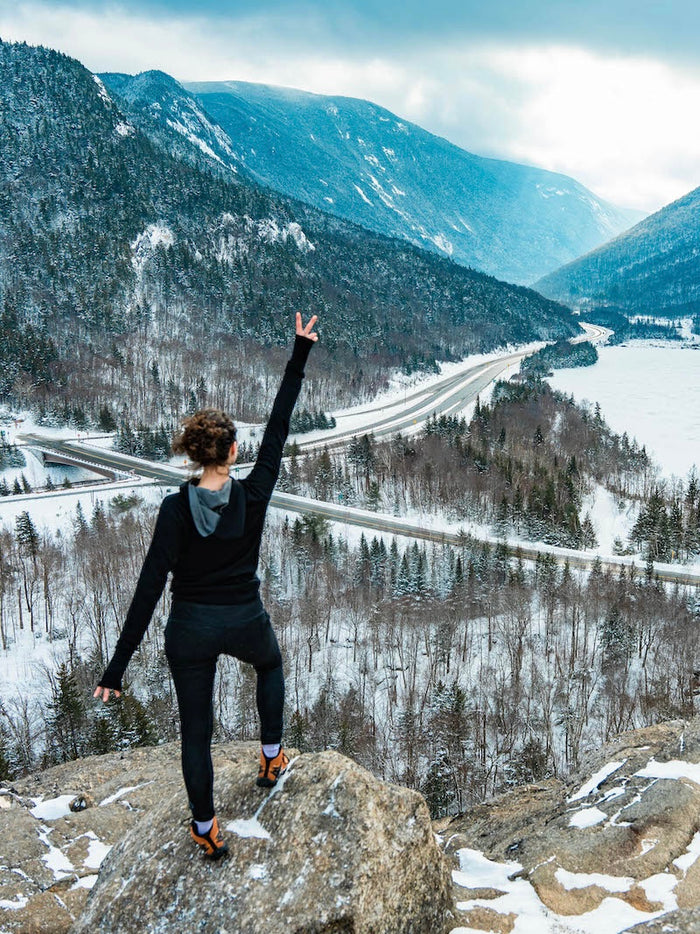 Woman Hiker in Lavender / Light Purple Alpaca Crew Socks Showing Peace Sign on Rocky Overlook of Snowy Valley and Roads – PAKA Apparel