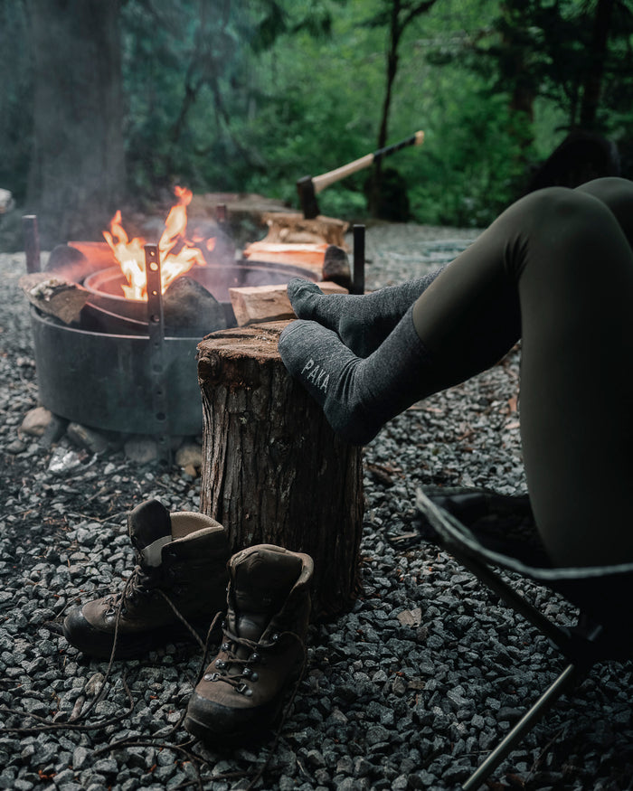 Dark Grey Sustainable Alpaca Wool Crew Socks on Feet Resting on Wood Stump in Front of Boots and Campfire – PAKA Apparel on Woman Model